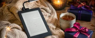 A cozy reading gift setup with an e-reader, a warm blanket, a book light, and a mug