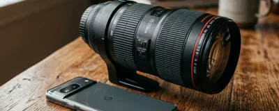 A sleek, modern smartphone lying flat on a wooden table with a massive, heavy professional DSLR camera lens absurdly attached to it.