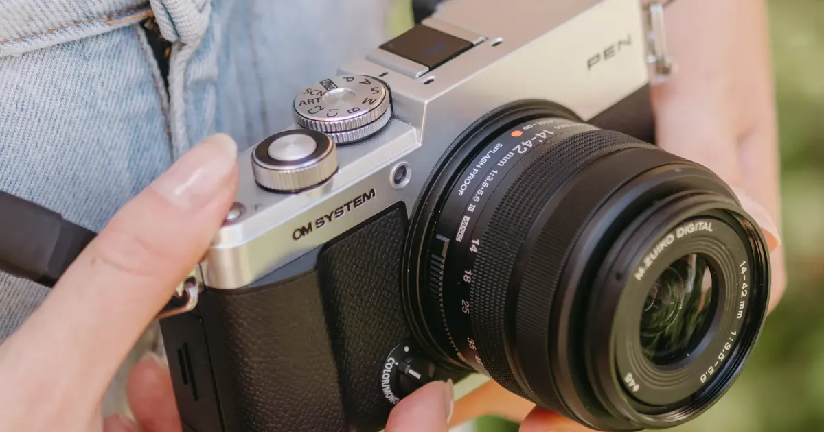 A retro-styled OM SYSTEM PEN mirrorless camera with silver metal dials resting on a wooden café table next to an espresso.