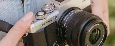 A retro-styled OM SYSTEM PEN mirrorless camera with silver metal dials resting on a wooden café table next to an espresso.