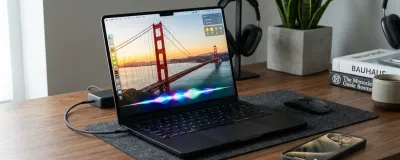 A Space Black MacBook Pro running macOS Golden Gate and an iPhone with glowing Siri AI waveforms on a wooden desk.