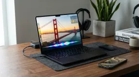 A Space Black MacBook Pro running macOS Golden Gate and an iPhone with glowing Siri AI waveforms on a wooden desk.