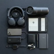A stylish and organized flat lay of modern commuting gadgets including noise-canceling headphones, a power bank, a sleek e-reader, and a smart travel mug, on a dark minimalist background