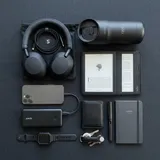 A stylish and organized flat lay of modern commuting gadgets including noise-canceling headphones, a power bank, a sleek e-reader, and a smart travel mug, on a dark minimalist background
