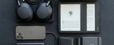 A stylish and organized flat lay of modern commuting gadgets including noise-canceling headphones, a power bank, a sleek e-reader, and a smart travel mug, on a dark minimalist background