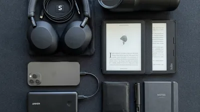 A stylish and organized flat lay of modern commuting gadgets including noise-canceling headphones, a power bank, a sleek e-reader, and a smart travel mug, on a dark minimalist background