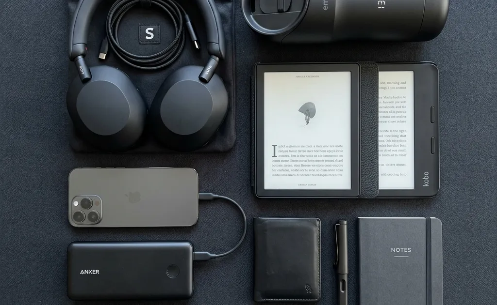 A stylish and organized flat lay of modern commuting gadgets including noise-canceling headphones, a power bank, a sleek e-reader, and a smart travel mug, on a dark minimalist background