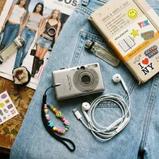 A vintage 2000s compact digital camera and wired Apple EarPods on a denim background, representing the Gen Z retro tech trend