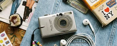 A vintage 2000s compact digital camera and wired Apple EarPods on a denim background, representing the Gen Z retro tech trend