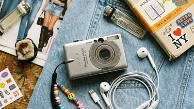 A vintage 2000s compact digital camera and wired Apple EarPods on a denim background, representing the Gen Z retro tech trend