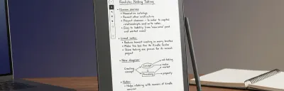 Kindle Scribe on an office desk with handwritten meeting notes on screen next to a laptop.